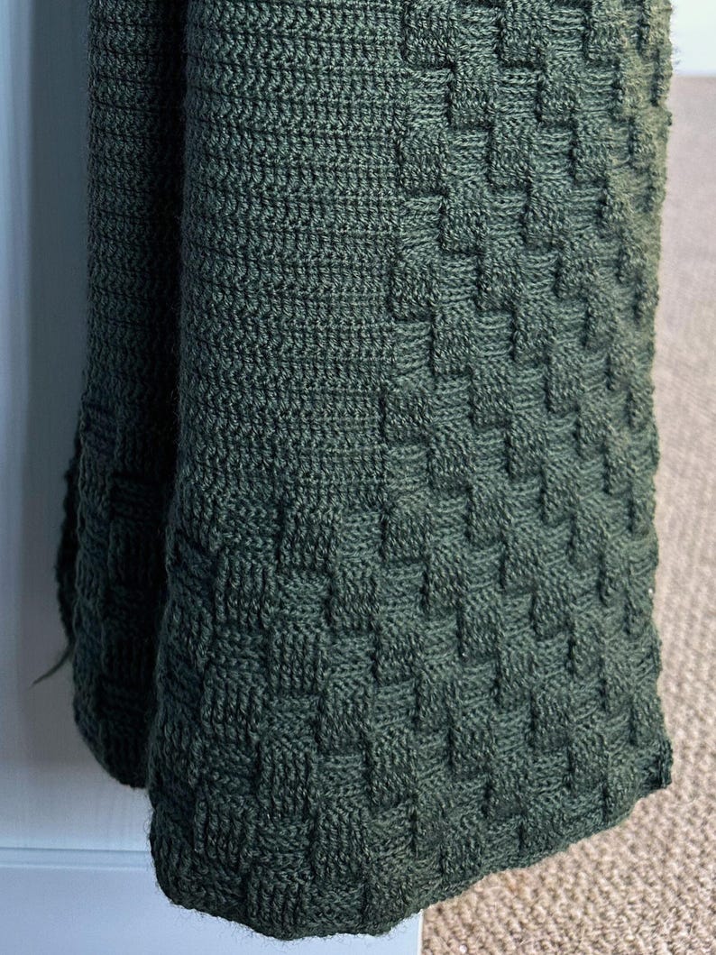 May include: A close-up of a dark green knitted blanket with a woven basket weave pattern. The blanket is folded over, revealing the texture and depth of the knit. The color is a deep, earthy green.