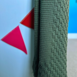 May include: A folded, olive green knitted blanket with a textured, basketweave pattern. The blanket is hanging, revealing its woven design and color. The background includes a white surface with red and pink triangle decorations and a blue wall.