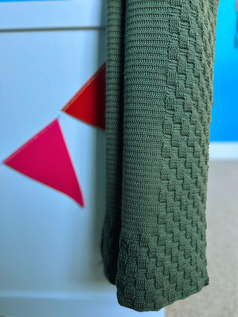 May include: A folded, olive green knitted blanket with a textured, basketweave pattern. The blanket is hanging, revealing its woven design and color. The background includes a white surface with red and pink triangle decorations and a blue wall.
