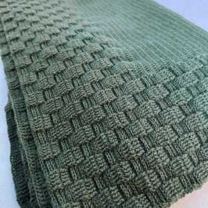 May include: A folded, sage green knitted blanket with a textured basketweave pattern. The blanket is made of soft material and is folded neatly, showcasing the detailed weave. The color is a muted, earthy green.