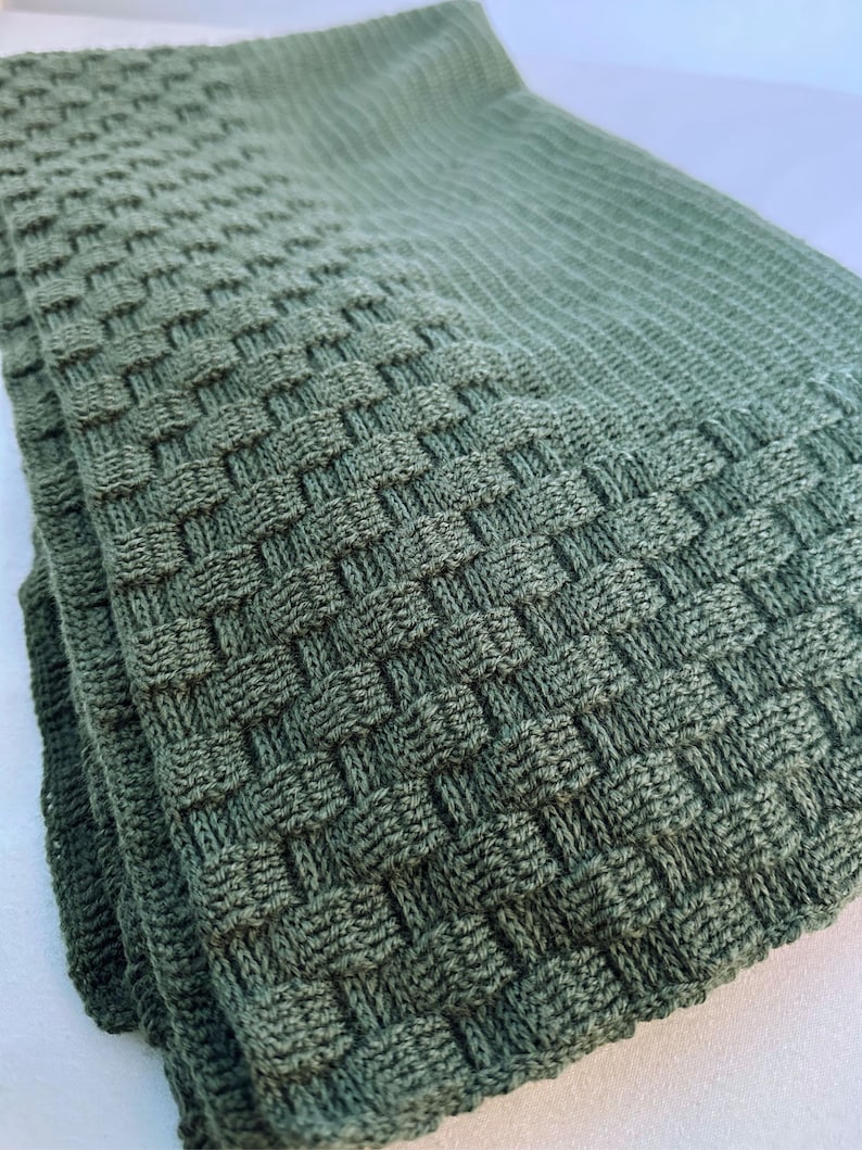 May include: A folded, sage green knitted blanket with a textured basketweave pattern. The blanket is made of soft material and is folded neatly, showcasing the detailed weave. The color is a muted, earthy green.