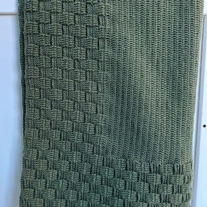 May include: A folded, olive green knitted blanket. The blanket features a woven basket-like pattern on the bottom third and a ribbed pattern on the upper two-thirds. The blanket is draped over a white surface.