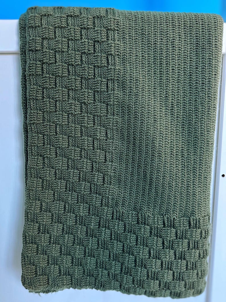 May include: A folded, olive green knitted blanket. The blanket features a woven basket-like pattern on the bottom third and a ribbed pattern on the upper two-thirds. The blanket is draped over a white surface.