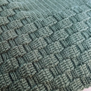 May include: Close-up of a woven, textured blanket in shades of green. The blanket features a basketweave pattern, with raised squares and a soft, inviting appearance. The knitted fabric is tightly woven, creating a cozy and durable textile.