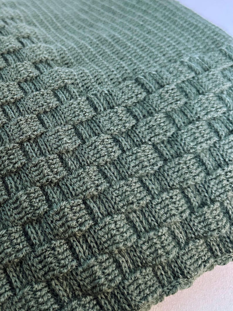 May include: Close-up of a woven, textured blanket in shades of green. The blanket features a basketweave pattern, with raised squares and a soft, inviting appearance. The knitted fabric is tightly woven, creating a cozy and durable textile.