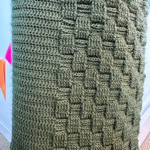 May include: Close-up of a knitted blanket in a muted olive green color. The blanket features a textured pattern with a woven basket design. The knitted material is soft and appears to be made of wool or a similar fiber.