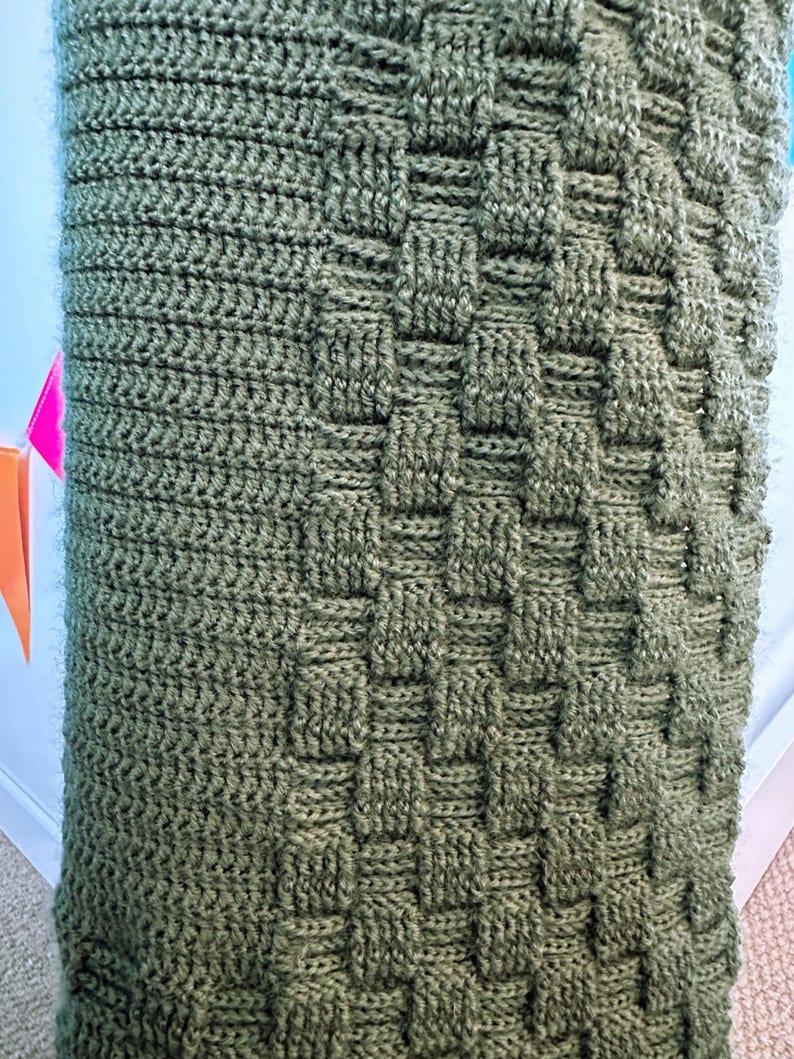May include: Close-up of a knitted blanket in a muted olive green color. The blanket features a textured pattern with a woven basket design. The knitted material is soft and appears to be made of wool or a similar fiber.