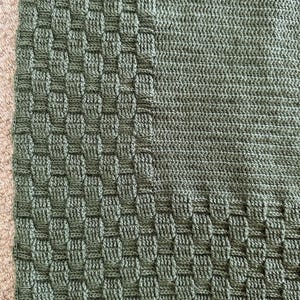 May include: Close-up of a green knitted blanket with a textured, woven-like border. The blanket's main body has a smooth, ribbed texture. The color is a muted, earthy green, and the blanket appears soft and cozy.