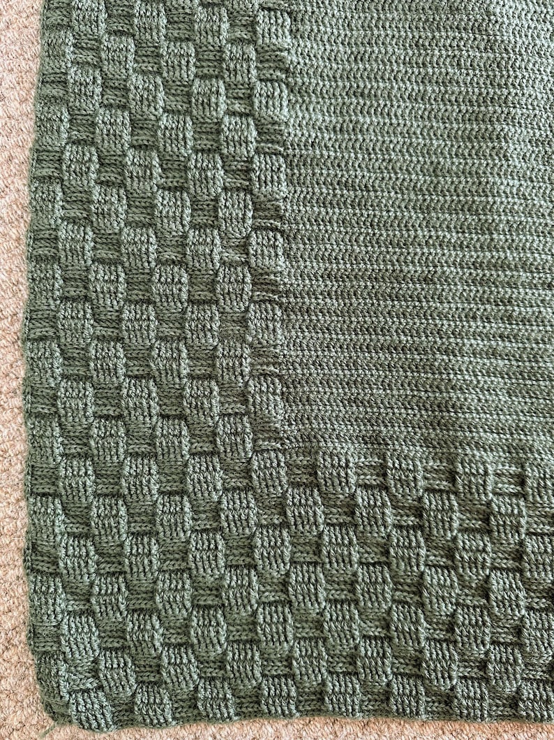 May include: Close-up of a green knitted blanket with a textured, woven-like border. The blanket's main body has a smooth, ribbed texture. The color is a muted, earthy green, and the blanket appears soft and cozy.