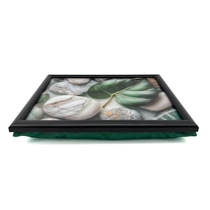 May include: A rectangular lap tray featuring a black frame and a printed surface depicting smooth white marble stones and a large green leaf. The base of the lap tray is made of a solid dark green fabric cushion, providing a stable surface for use.