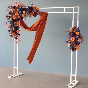 May include: A white metal wedding arch adorned with vibrant flowers in orange, blue, and red hues. An orange fabric drape adds a decorative element. The arch is set against a pale blue backdrop.