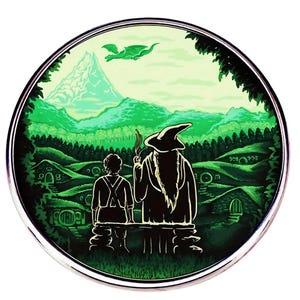 May include: Circular green and white illustration featuring two figures overlooking a landscape with rolling hills, hobbit holes, and a mountain. A dragon flies in the sky. The scene is framed by a silver border.