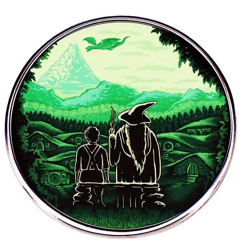 May include: Circular green and white illustration featuring two figures overlooking a landscape with rolling hills, hobbit holes, and a mountain. A dragon flies in the sky. The scene is framed by a silver border.
