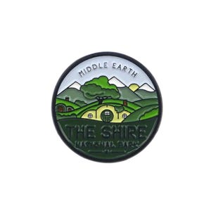 May include: A round enamel pin featuring a landscape of The Shire from Middle Earth. The pin has a black border and depicts rolling green hills, hobbit holes, and the text "THE SHIRE" and "MIDDLE EARTH".