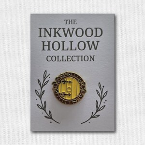 May include: A pin featuring a yellow door design with a brown frame and accents, set against a gray card. The card reads "THE INKWOOD HOLLOW COLLECTION" in dark gray text, with leaf illustrations. A decorative accessory.