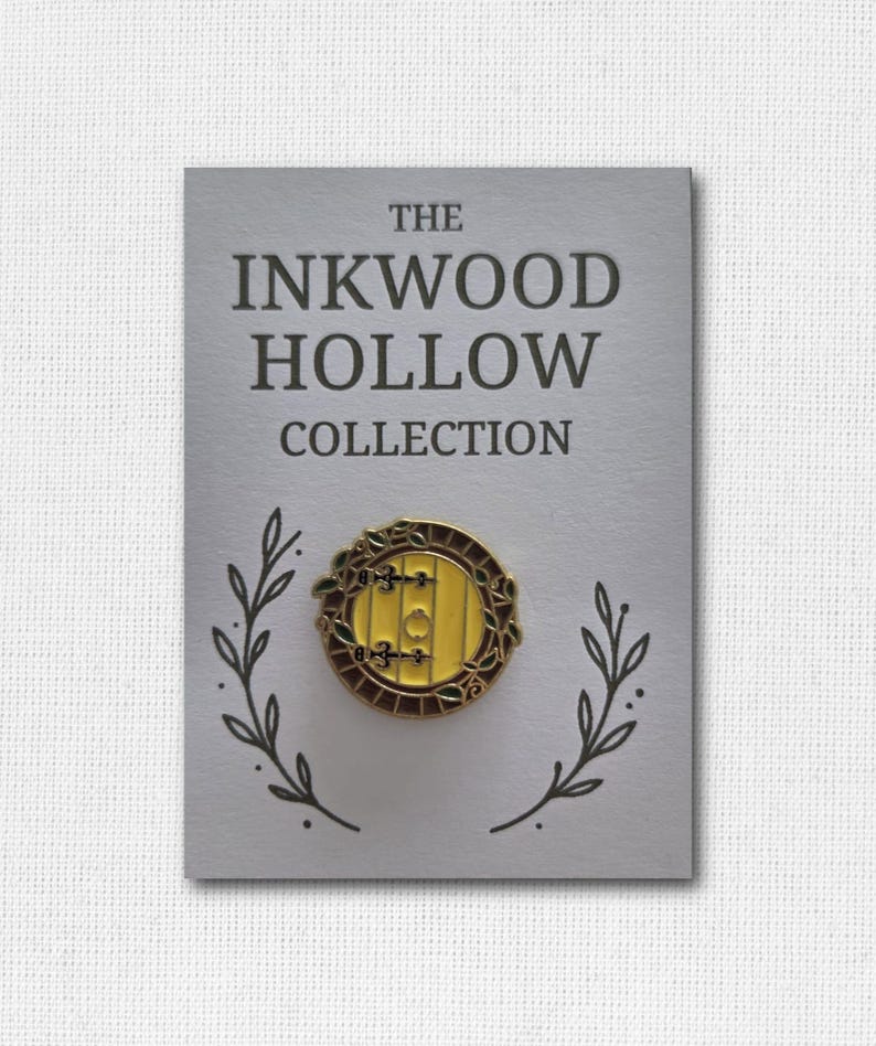 May include: A pin featuring a yellow door design with a brown frame and accents, set against a gray card. The card reads "THE INKWOOD HOLLOW COLLECTION" in dark gray text, with leaf illustrations. A decorative accessory.