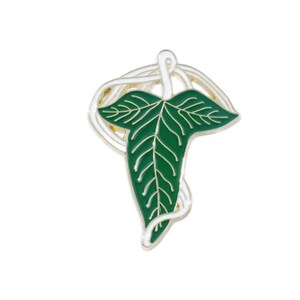 May include: A green leaf-shaped enamel pin with white veins and a silver-colored border. The pin features a stylized leaf design with a swirling, decorative element above the leaf. The pin is set against a white background.
