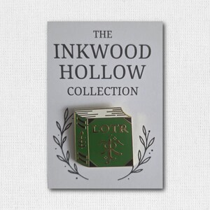 May include: A green enamel pin in the shape of a book with the letters "LOTR" on the cover. The pin is on a white card with the text "THE INKWOOD HOLLOW COLLECTION" and a decorative leaf design.