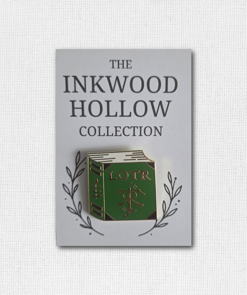 May include: A green enamel pin in the shape of a book with the letters "LOTR" on the cover. The pin is on a white card with the text "THE INKWOOD HOLLOW COLLECTION" and a decorative leaf design.