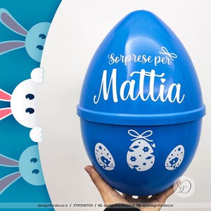 Könnte beinhalten: Ein blauer, eiförmiger Kunststoffbehälter mit dem Text "Sorprese per Mattia" in weißer Schrift. Das Ei hat drei dekorative Eierdesigns und wird von einer Hand gehalten. Der Hintergrund zeigt Hasen-Grafiken.