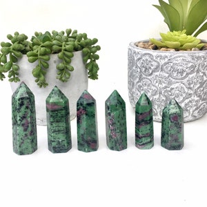 May include: Seven green and pink zultanite crystal points arranged in a row on a white surface. The crystals are polished and have a smooth, glassy finish.