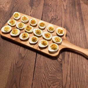 May include: A wooden cutting board with a handle, filled with deviled eggs sprinkled with paprika.