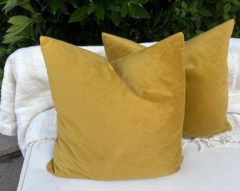 May include: Two square throw pillows made of a solid mustard yellow velvet fabric. The pillows have a smooth, soft texture and are positioned upright, showcasing a consistent, deep yellow color across their entire visible surface area.