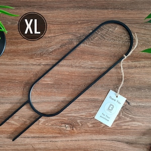 Pode incluir: Um suporte para plantas de metal preto com uma forma oval grande e duas pernas. O suporte está etiquetado como "XL" e está sobre uma superfície de madeira.