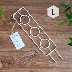 Op de afbeelding: Een witte metalen plantenstandaard met drie ronde openingen. De standaard is ontworpen om drie kleine potplanten te houden. De standaard is gelabeld met een "L" voor de maat.