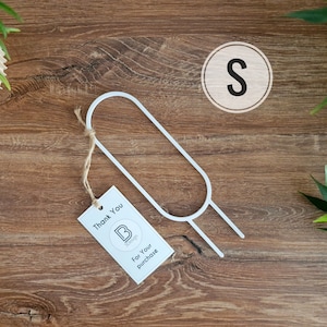 Könnte beinhalten: Ein weißer Metall-Kleiderbügel mit einem Etikett mit der Aufschrift "Thank You For Your Purchase". Ein Holzkreis mit dem Buchstaben "S" ist ebenfalls sichtbar. Die Artikel befinden sich auf einer Holzoberfläche mit einer Pflanze im Hintergrund.