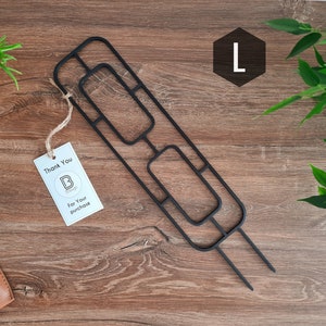 Può includere: Portavaso in metallo nero con un design geometrico. Il portavaso ha tre aperture rettangolari e due punte lunghe per l'inserimento nel terreno. Il portavaso è etichettato con la lettera "L" in un esagono.