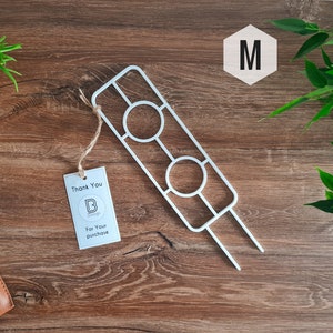 May include: A white metal plant support with two circular cutouts and a tag that says "Thank You". The support is on a wooden surface with greenery and a wooden "M" in the background. The tag says "For Your purchase".