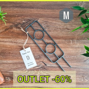 Op de afbeelding: Een zwarte metalen plantenstandaard met twee ronde openingen. De standaard staat op een houten oppervlak met een bruine leren hoes en een klein wit label met de tekst "Bedankt" en een logo. De tekst "OUTLET -60%" is zichtbaar in de rechterbenedenhoek van de afbeelding. De letter "M" is zichtbaar in de rechterbovenhoek van de afbeelding.