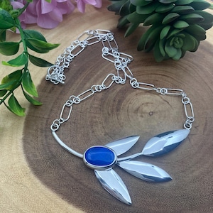 May include: A silver necklace with a leaf design. The necklace features a large oval blue stone in the center, surrounded by three silver leaf-shaped pendants. The chain is made of interconnected silver links.