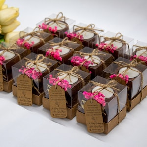 May include: A collection of small, clear boxes filled with brown coconut shell candle holders. Each holder has a small white candle and pink dried flowers. The boxes are tied with twine and have a brown paper tag with the text "In Loving Memory" and a date.