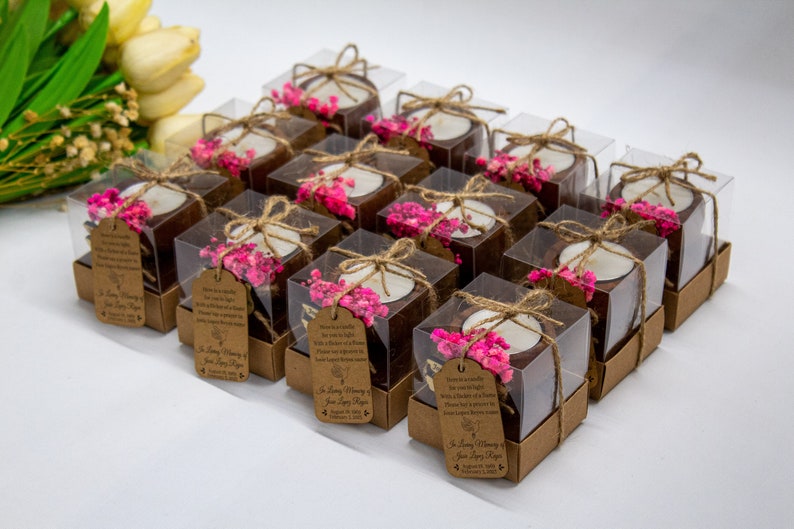 May include: A collection of small, clear boxes filled with brown coconut shell candle holders. Each holder has a small white candle and pink dried flowers. The boxes are tied with twine and have a brown paper tag with the text "In Loving Memory" and a date.