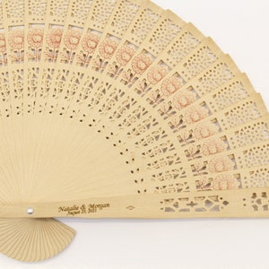May include: A light brown wooden hand fan with a delicate floral design. The fan is closed and has a handle with the text "Natalie & Morgan August 24, 2017" engraved on it.