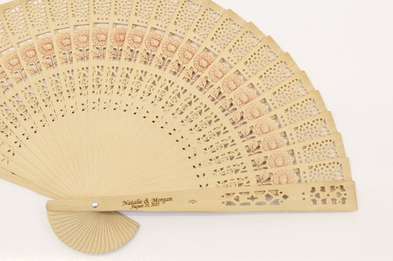 May include: A light brown wooden hand fan with a delicate floral design. The fan is closed and has a handle with the text "Natalie & Morgan August 24, 2017" engraved on it.