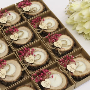 May include: A set of small, rustic wedding favors. Each favor is a wooden slice with a small candle in the center. The wooden slices are decorated with a small heart-shaped tag with the names "Julia & Joseph" and the wedding date. The favors are packaged in small, brown boxes with pink flowers.