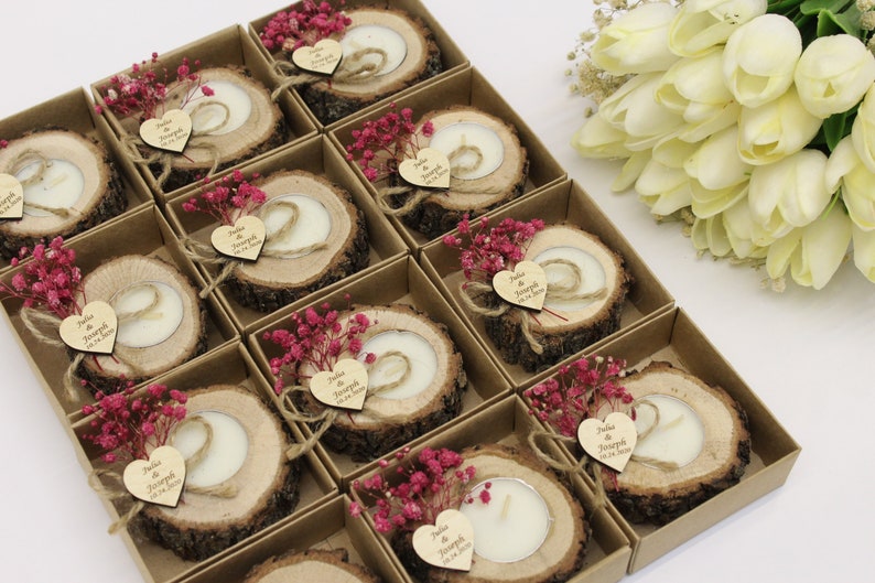 May include: A set of small, rustic wedding favors. Each favor is a wooden slice with a small candle in the center. The wooden slices are decorated with a small heart-shaped tag with the names "Julia & Joseph" and the wedding date. The favors are packaged in small, brown boxes with pink flowers.