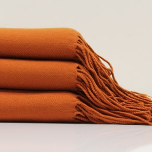 May include: A stack of three orange throw blankets with fringe edges. The blankets are folded and arranged on a white surface.