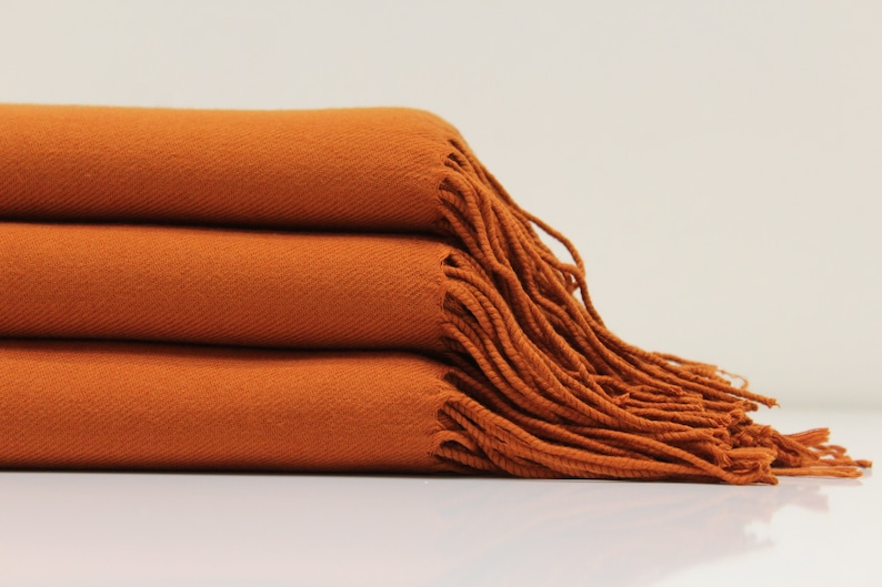 May include: A stack of three orange throw blankets with fringe edges. The blankets are folded and arranged on a white surface.