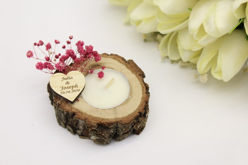 May include: A small wooden heart with the text "Julia & Joseph 10.24.2020" sits on a wooden slice with a small white candle in the center. Pink dried flowers decorate the arrangement.