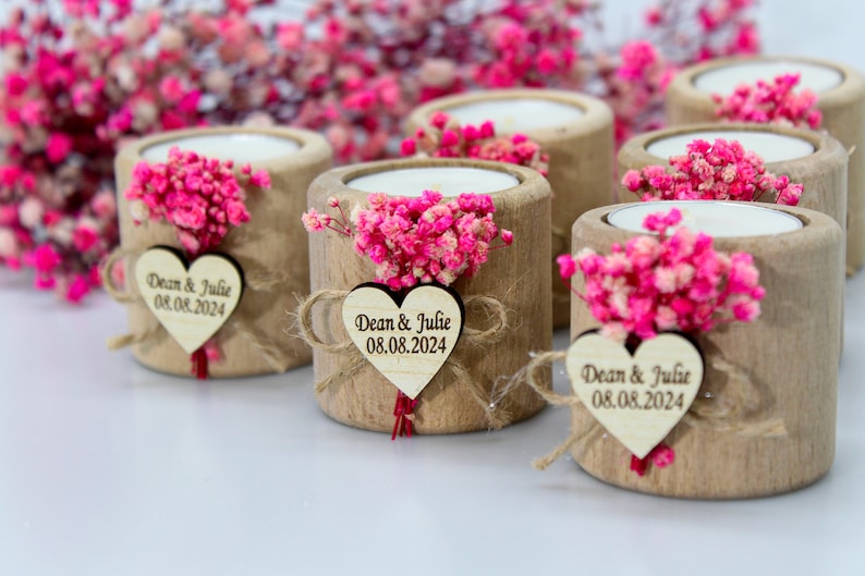 May include: Wooden candle holders with a small bouquet of pink flowers and a heart-shaped tag with the text "Dean & Julie 08.08.2024" on each.