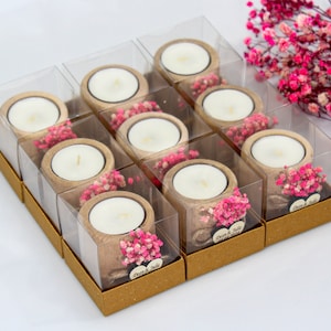 May include: A set of 10 small, clear glass candle holders with a wooden base and pink dried flowers. Each holder has a small white candle inside and a brown paper box with the text "Dear & Julie" printed on it.