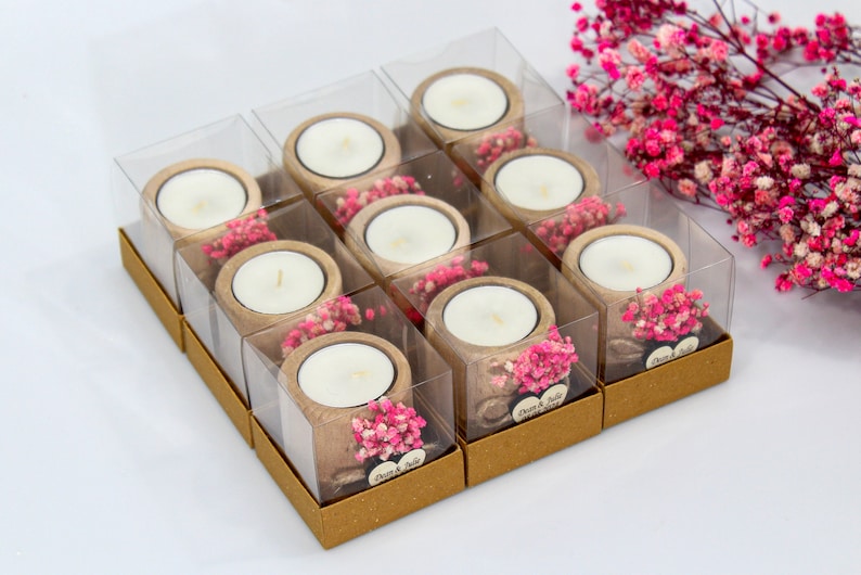 May include: A set of 10 small, clear glass candle holders with a wooden base and pink dried flowers. Each holder has a small white candle inside and a brown paper box with the text "Dear & Julie" printed on it.