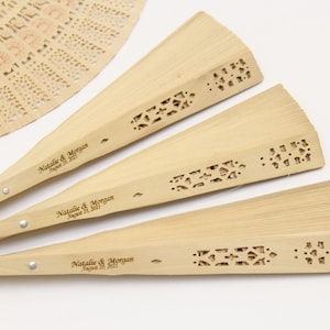 May include: Three wooden hand fans with a decorative cut-out design and personalised with the text "Natalie & Morgan August 28, 2021".
