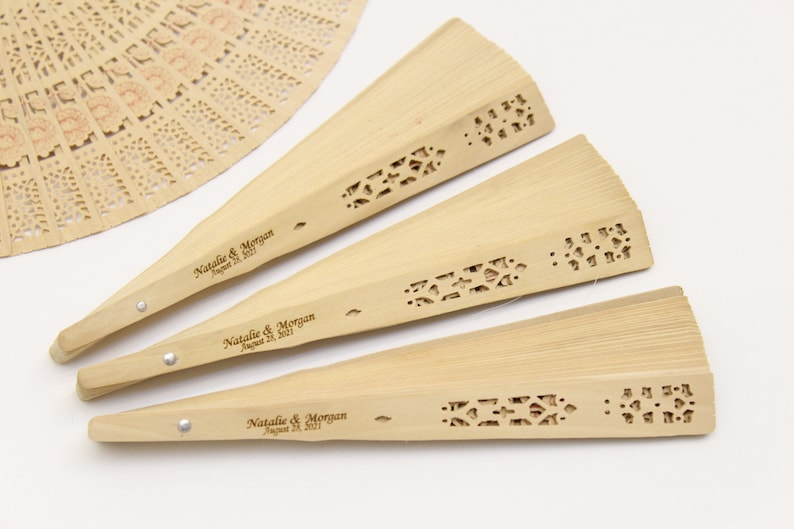 May include: Three wooden hand fans with a decorative cut-out design and personalised with the text "Natalie & Morgan August 28, 2021".