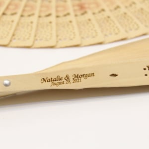 May include: A wooden hand fan with a carved design and the text "Natalie & Morgan August 28, 2021" engraved on the handle.