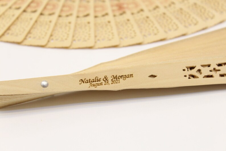 May include: A wooden hand fan with a carved design and the text "Natalie & Morgan August 28, 2021" engraved on the handle.
